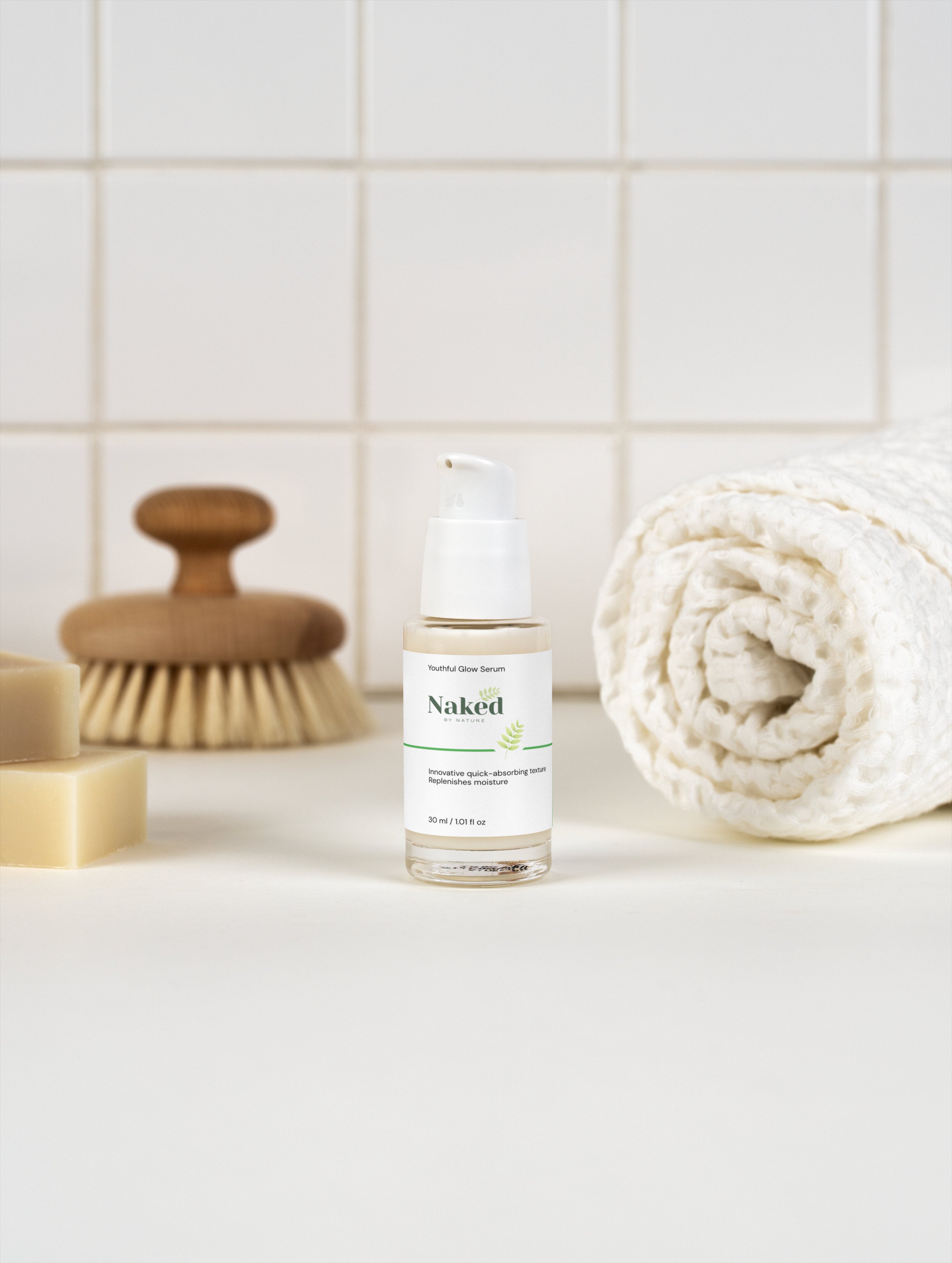 Youthful Glow Serum in clear bottle with pump, beside wooden brush and rolled towel on white tile background