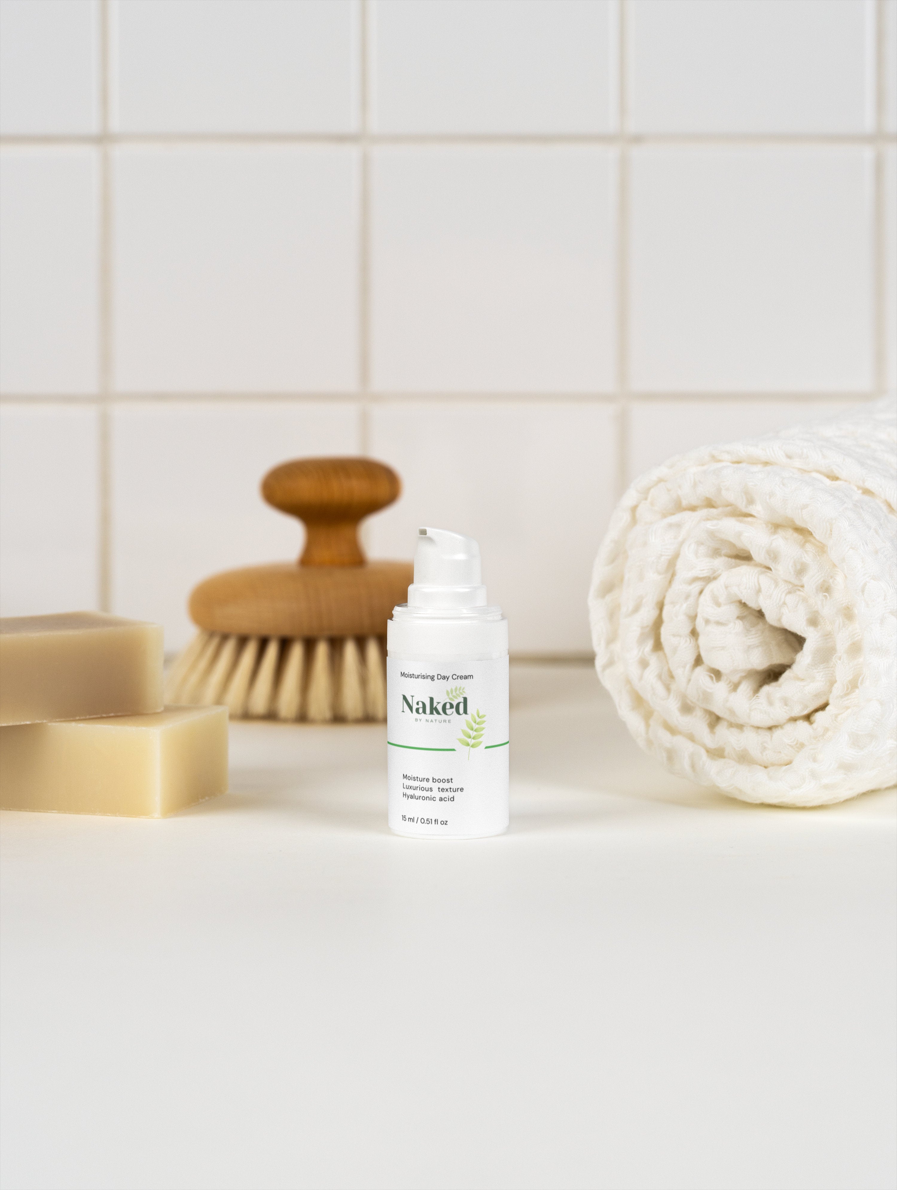 Naked by Nature Moisturising Day Cream with hyaluronic acid, beside soap bars, wooden brush, and rolled towel on tiled surface